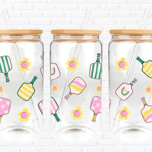 May include: Three clear glass drinking jars with bamboo lids and clear straws. Each jar is decorated with colourful pickleball paddles and yellow flowers. The paddles have green, pink, and yellow designs. The jars are set against a white brick wall.