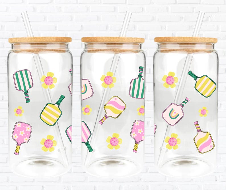 May include: Three clear glass drinking jars with bamboo lids and clear straws. Each jar is decorated with colourful pickleball paddles and yellow flowers. The paddles have green, pink, and yellow designs. The jars are set against a white brick wall.