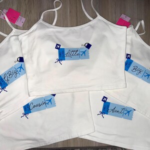 Puede incluir: Camisetas sin mangas blancas con tirantes finos, cada una con un gráfico azul y blanco de un billete de avión. Los billetes tienen el texto "Little", "Big", "Cousin" y "Aunt". Las camisetas son para un evento familiar.