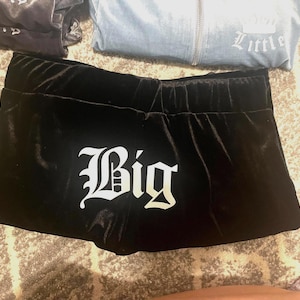 Puede incluir: Pantalones cortos de terciopelo negro con la palabra "Big" en letras góticas blancas. Los pantalones cortos tienen una cintura elástica. Otras prendas de vestir son visibles en el fondo.