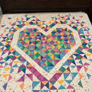 May include: A colorful quilt with a large heart design in the center. The heart is made up of many small triangles in shades of pink, purple, blue, green, yellow, and orange. The quilt is surrounded by a white border with a small floral pattern.