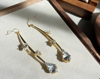 May include: A pair of gold earrings with clear crystal stones. The earrings are long and dangle, with the stones arranged in a geometric pattern.