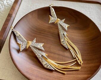 May include: A pair of silver and gold earrings with a bow and tassel design. The earrings are set with clear rhinestones and have a dangling tassel of gold chains.