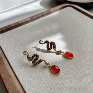 May include: A pair of gold snake earrings with red gemstone accents. The earrings are dangling and feature a snake design with a red gemstone at the end of each earring.