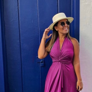 Może przedstawiać: Kobieta w fioletowej sukience z dekoltem halterowym i węzłem w talii, białym kapeluszu i brązowych sandałach. Stoi przed niebieskimi drzwiami.