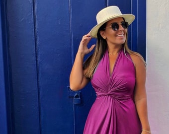 May include: A woman wearing a purple halter dress with a knot at the waist, a white hat, and brown sandals. She is standing in front of a blue door.