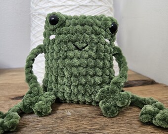 Op de afbeelding: Een kleine, olijfgroene gehaakte kikkerknuffel met een vierkant lichaam, lange dunne armen en grote zwarte plastic ogen. De kikker heeft een eenvoudig geborduurde zwarte mond en twee kleine witte stiksels onder de ogen.