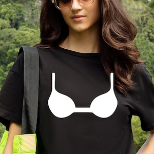 Op de afbeelding: Zwart T-shirt met een witte bh-afbeelding. Het shirt heeft een ronde hals. Het model draagt een zonnebril en heeft lang bruin haar. De achtergrond toont een wazig beeld van groene bomen.