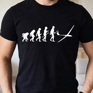 May include: A black short-sleeved t-shirt featuring a white graphic print that depicts the evolution of humans, starting from a primate and progressing to a modern human figure holding a glider aircraft, which is positioned at the end of the sequence.