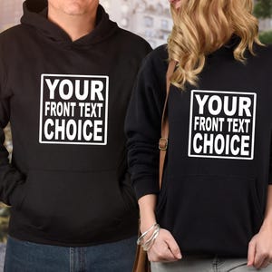Puede incluir: Sudaderas con capucha negras con un diseño rectangular blanco en la parte delantera que dice "YOUR FRONT TEXT CHOICE". Las sudaderas tienen un bolsillo delantero y las usan dos personas. El fondo está borroso.