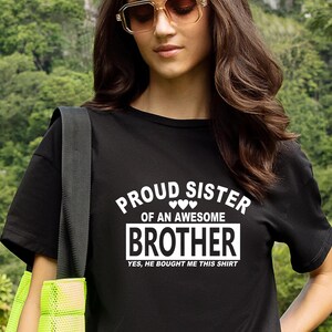 May include: Black t-shirt with the text "PROUD SISTER OF AN AWESOME BROTHER" in white. The text "YES, HE BOUGHT ME THIS SHIRT" is below the word "BROTHER". The person is wearing sunglasses and has long brown hair.