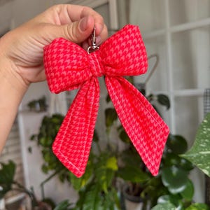 Puede incluir: Un llavero rojo en forma de lazo con un estampado de pata de gallo. El lazo es de tela y tiene un cierre plateado. El lazo es de color rojo brillante con un patr&oacute;n rojo m&aacute;s claro.