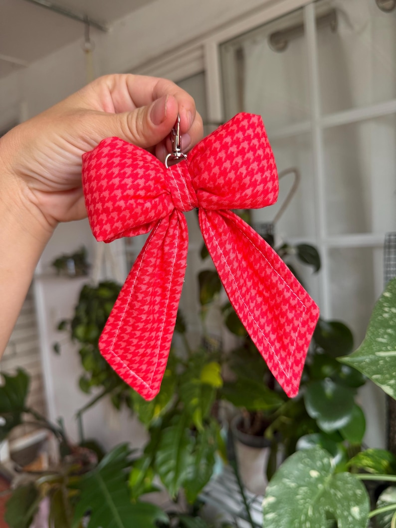 Puede incluir: Un llavero rojo en forma de lazo con un estampado de pata de gallo. El lazo es de tela y tiene un cierre plateado. El lazo es de color rojo brillante con un patr&oacute;n rojo m&aacute;s claro.