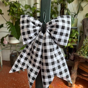 May include: A large decorative bow featuring a black and white buffalo check pattern. The bow is constructed with two wide top loops and two long, pointed tails hanging downward, secured in the center by a solid black fabric band.