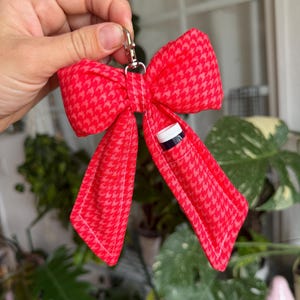 Puede incluir: Un llavero rojo en forma de lazo con un estampado de pata de gallo. El lazo tiene un peque&ntilde;o bolsillo que contiene una botella peque&ntilde;a con una tapa blanca. Un cierre de color plateado est&aacute; unido a la parte superior del lazo.