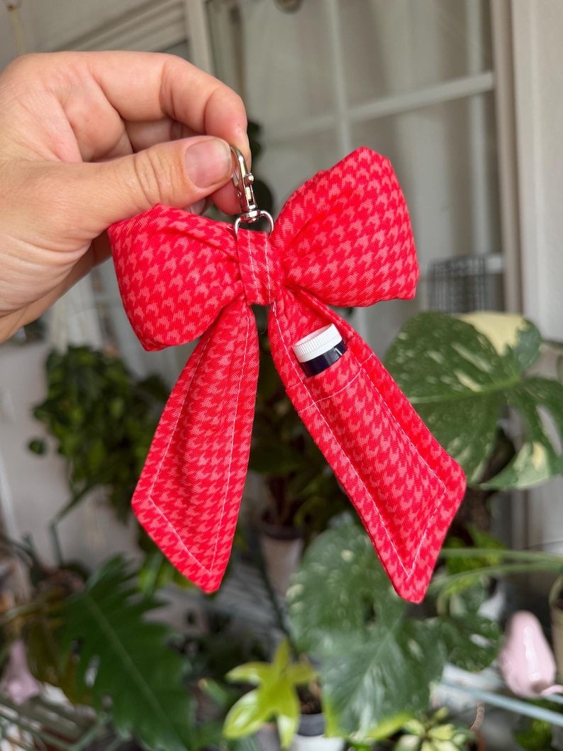 Puede incluir: Un llavero rojo en forma de lazo con un estampado de pata de gallo. El lazo tiene un peque&ntilde;o bolsillo que contiene una botella peque&ntilde;a con una tapa blanca. Un cierre de color plateado est&aacute; unido a la parte superior del lazo.