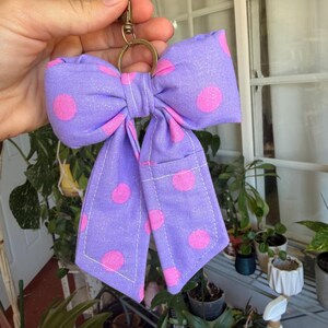 Puede incluir: Un llavero hecho a mano con forma de lazo morado con lunares rosas. El lazo es de tela y tiene un cierre y un anillo de metal dorado. El lazo mide aproximadamente 13 cm de ancho.
