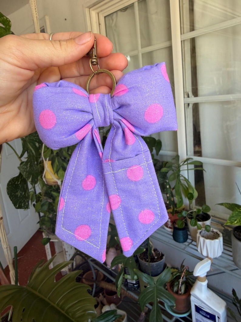 Puede incluir: Un llavero hecho a mano con forma de lazo morado con lunares rosas. El lazo es de tela y tiene un cierre y un anillo de metal dorado. El lazo mide aproximadamente 13 cm de ancho.