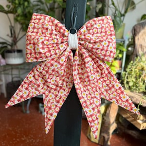May include: A large decorative bow made of fabric featuring a red and white gingham check pattern overlaid with a small white daisy print. The bow is gathered at the center with a white fabric band and hangs from a black metal hook.