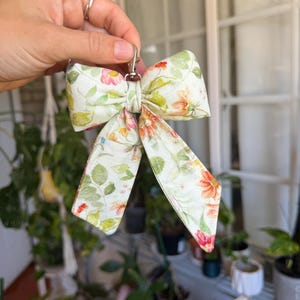 Puede incluir: Un llavero con lazo estampado floral y cierre plateado. El lazo presenta un patr&oacute;n de hojas verdes y flores naranjas, rosas y rojas sobre un fondo crema. El lazo est&aacute; sostenido por una mano.