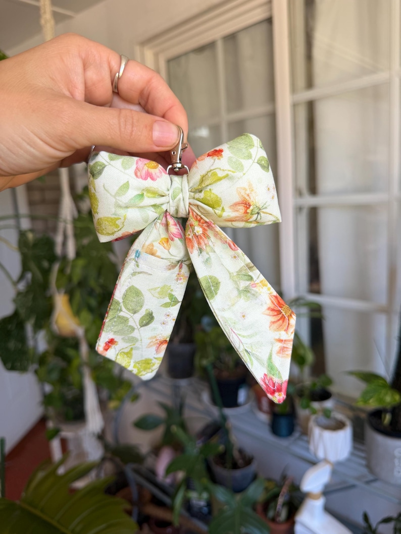Puede incluir: Un llavero con lazo estampado floral y cierre plateado. El lazo presenta un patr&oacute;n de hojas verdes y flores naranjas, rosas y rojas sobre un fondo crema. El lazo est&aacute; sostenido por una mano.