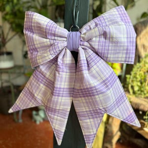 May include: A large decorative bow made of fabric with a light purple and white plaid pattern. The bow features two wide top loops and two pointed tails hanging downward, secured in the center by a solid light purple fabric band and a metal ring.