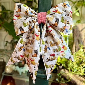 May include: A large, decorative bow featuring a white fabric with a repeating pattern of various dog breeds. The bow is tied with a red and white gingham ribbon. The bow is attached to a green post, with greenery in the background.