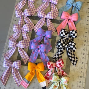 Puede incluir: Una colecci&oacute;n de lazos de tela hechos a mano en varios patrones y colores, incluyendo cuadros, lunares e estampados florales. Algunos lazos son de colores lisos, mientras que otros presentan un dise&ntilde;o de l&aacute;piz o estampado de cerezas. Cada lazo tiene un clip de metal.