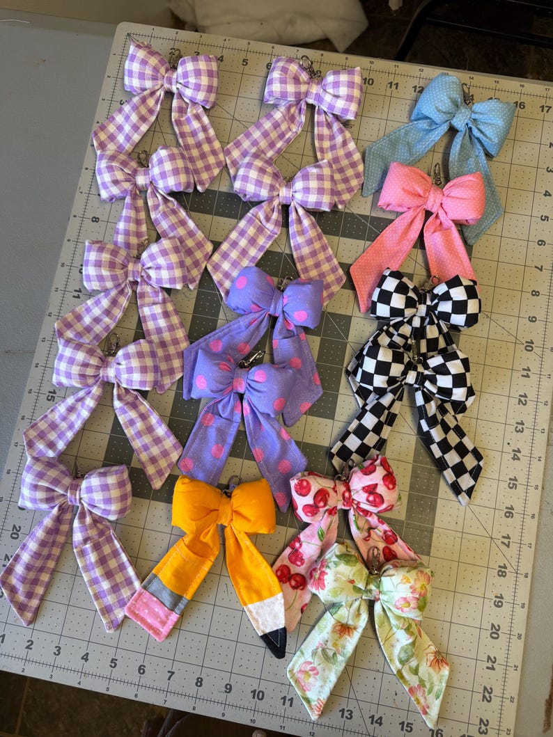 Puede incluir: Una colecci&oacute;n de lazos de tela hechos a mano en varios patrones y colores, incluyendo cuadros, lunares e estampados florales. Algunos lazos son de colores lisos, mientras que otros presentan un dise&ntilde;o de l&aacute;piz o estampado de cerezas. Cada lazo tiene un clip de metal.
