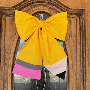 Puede incluir: Un gran lazo decorativo amarillo de tela con forma de lápiz, colgado en una puerta de madera. El diseño del lápiz incluye detalles grises, rosas, blancos y negros. El lazo está anudado en el centro y tiene extremos largos.