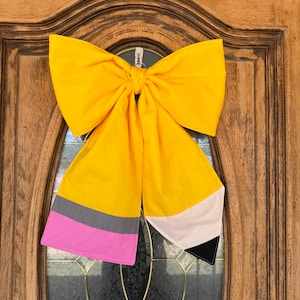 May include: A large, yellow fabric bow shaped like a pencil, hanging on a wooden door. The pencil design features gray, pink, white, and black accents. The bow is tied in the center and has long, flowing ends, creating a whimsical school-themed decoration.