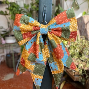 May include: A large fabric bow featuring a patchwork design of squares in orange, olive green, and light blue. The fabric is decorated with small white polka dots and tiny yellow flower patterns. The bow is secured with a light blue center knot.