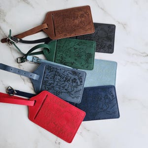 May include: A collection of leather luggage tags in various colors, including brown, green, blue, and red. Each tag features a detailed map design and a strap with a buckle for attachment. The tags are arranged on a marble surface.