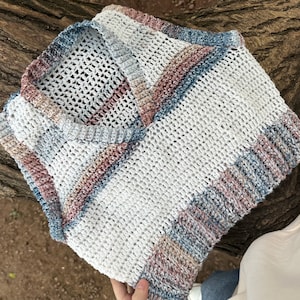 Può includere: Un top all'uncinetto bianco fatto a mano con scollatura quadrata. Il top presenta strisce di filato blu, rosa e beige. Il design a trama aperta è visibile. Il top è piegato e appoggiato su una superficie di legno.