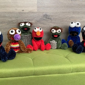 May include: A collection of seven handmade crochet character dolls, including Elmo, Cookie Monster, and Grover, in various colors. The dolls are seated on a green upholstered bench against a wooden background. Each doll has unique features.