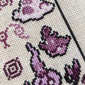 May include: Close-up of a cross-stitch pattern featuring a purple and white ghost design. The ghost is outlined in black and has a simple, pixelated design.