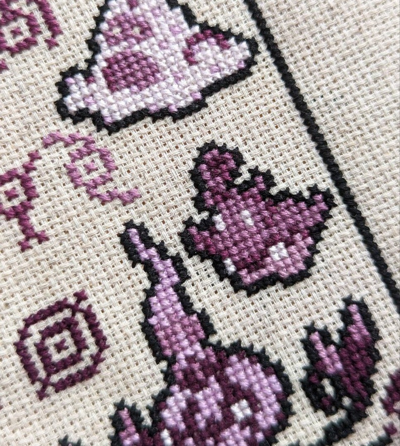 May include: Close-up of a cross-stitch pattern featuring a purple and white ghost design. The ghost is outlined in black and has a simple, pixelated design.