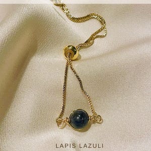 Op de afbeelding: Een goudkleurige verstelbare armband met een donkerblauwe lapis lazuli steen. De armband heeft een fijne ketting en een schuifkraal voor aanpassing. De tekst "MORE VARIATIONS" en "LAPIS LAZULI" zijn zichtbaar.