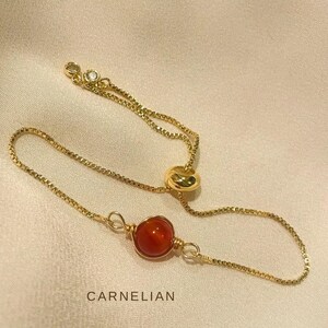 Op de afbeelding: Een goudkleurige verstelbare ring met een heldere oranje carneoolsteen. De ring heeft een delicaat kettingontwerp met een kleine, sprankelende accentsteen. De tekst "MORE VARIATIONS" en "CARNELIAN" zijn zichtbaar.