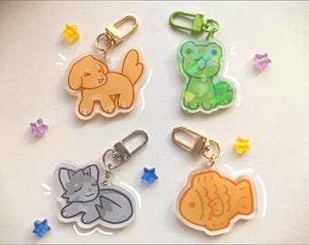 Puede incluir: Cuatro llaveros acrílicos con diseños de animales de dibujos animados: un perro, una rana, un gato y un pez. Cada llavero tiene un mosquetón de metal de diferente color. El perro es marrón, la rana es verde, el gato es gris y el pez es naranja.