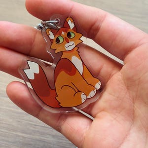 Puede incluir: Un llavero de acr&iacute;lico transparente con un gato de dibujos animados. El gato tiene pelaje naranja y blanco, ojos verdes y una expresi&oacute;n sonriente. Est&aacute; sentado con la cola enroscada. Un anillo de metal plateado est&aacute; unido en la parte superior.