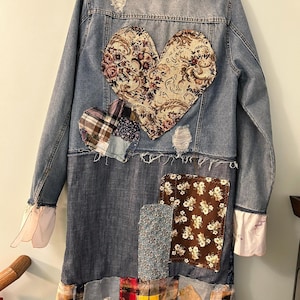 May include: A distressed denim jacket with patchwork details. The back features a large heart-shaped patch with floral fabric, and a smaller plaid heart. The jacket has a long, patchwork skirt and frayed edges.