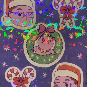 May include: A sticker sheet with a holographic background featuring a variety of holiday-themed illustrations. The sheet includes a person wearing a Santa hat, a wreath with a cat inside, and two candy canes with a bow. The text "Puppycat Holiday Sheet" and "Mysticaltoastart" are printed on the sheet.