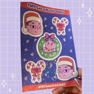 May include: A rectangular sticker sheet titled PUPPYCAT HOLIDAY SHEET with a dark blue background featuring white snowflakes. It includes two cartoon faces wearing Santa hats, two candy canes with red bows, and a pink creature in a green wreath.