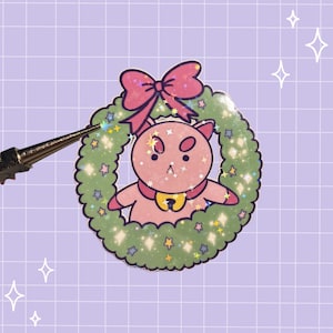 May include: A holographic sticker featuring a pink cat with a yellow collar centered inside a green wreath topped with a large pink bow. The design includes small colorful stars and a shimmering finish, held by a pair of metal tweezers.