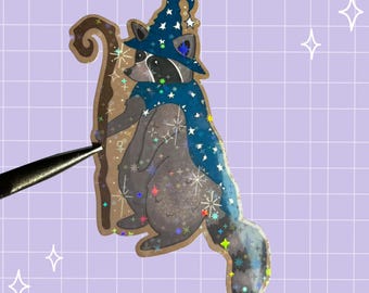 Könnte beinhalten: Ein holografischer Aufkleber eines Waschbären als Zauberer verkleidet, der einen blauen spitzen Hut und einen mit weißen Sternen verzierten Umhang trägt. Der Waschbär hält einen braunen Holzstab. Der Aufkleber hat ein schimmerndes Finish.