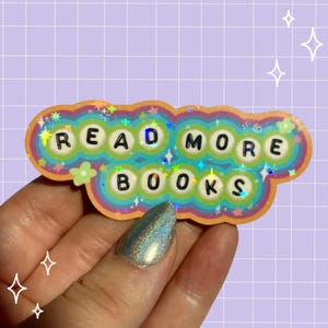 May include: A die-cut sticker featuring a rainbow-colored, wavy-edged background with a holographic finish. The sticker displays the text READ MORE BOOKS in black, rounded, bead-like letters, accented with small colorful stars and flower shapes.