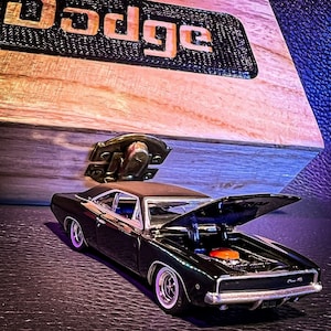 Puede incluir: Un coche modelo negro con el capó abierto, que revela un motor naranja, se encuentra frente a una caja de madera con el nombre "Dodge". El coche tiene neumáticos de banda blanca y detalles cromados. La caja tiene un cierre metálico.