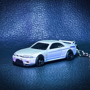 May include: A silver toy car with black wheels and a rear spoiler. The car has blue and white accents and a keychain attached. The car is on a dark blue surface.