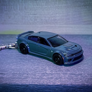 May include: A teal-colored toy car keychain with black wheels. The car has a detailed design, including a hood scoop and spoiler. The keychain is attached with a silver metal ring.